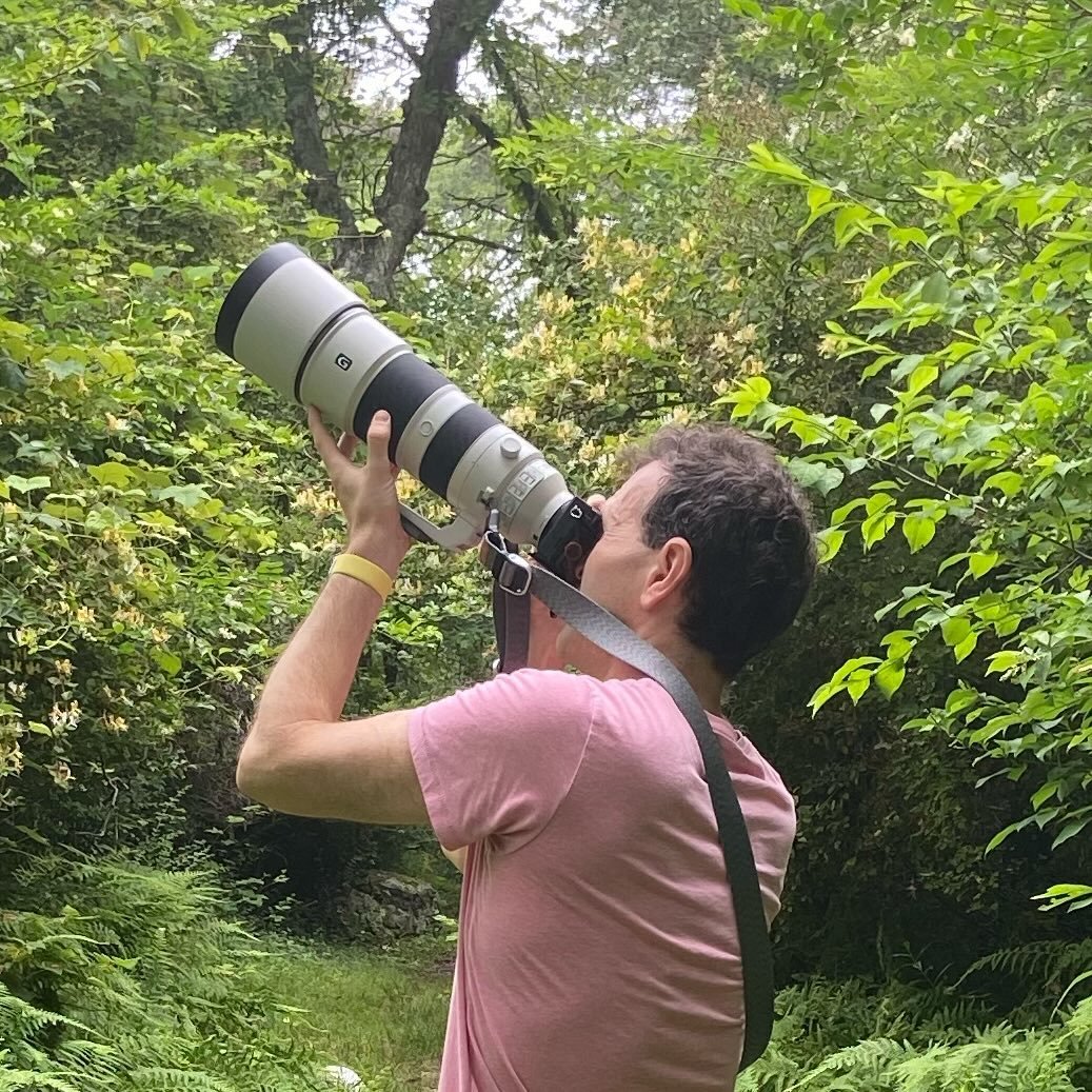 Keith in the woods pointing a large camera lens up into the sky